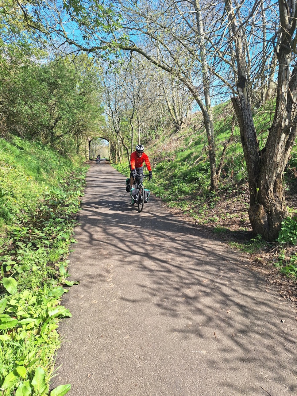 A man in an orange jumper and helmet cycles on a smooth surfaced path.