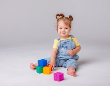 Phoebe is wearing a yellow t-shirt and blue dungarees with the word happy on. She is smiling and playing with five building blocks.