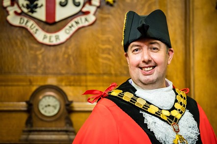 A picture of Cllr Troy Gallagher in Mayoral Robes standing in the Mayor's Parlour in Islington Town Hall in front of the borough crest