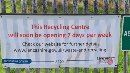 Household Waste Recycling Centre sign, on a railing, with grass behind. 
The white sign, in red font, reads: "This Recycling Centre will soon be opening 7 days per week."
In black: "check our website for further details
www.lancashire.gov.uk/waste-and-recycling"
On blue banner at bottom left: lancas