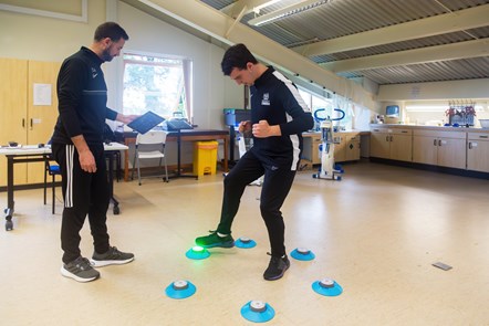 Two males in black and white sport training clothing, one on right doing a reaction speed test with his feet tapping light pods on the floor