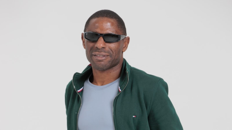 Darren Harris: Darren stands against a white background and smiles at the camera. He is a black man with short black hair and wears dark black sunglasses