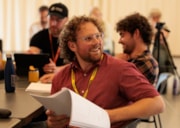 Sanctuary Queer Arts CIC: Sanctuary Queer Arts' Lead Artist, Jonathan O'Neill, looks to his left and smiles while holding a script.