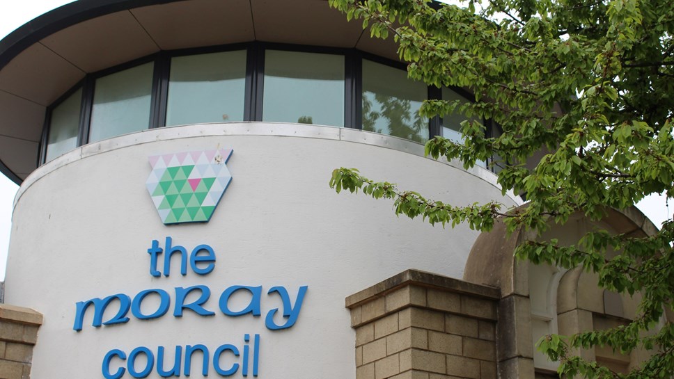 The image shows the front of the Moray Council headquarters building, featuring the council's name in blue letters on a light colored wall.