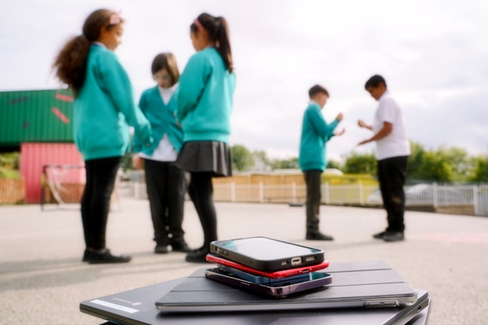 DisconnectToReconnectF: Image shows a close-up of devices, such as phones and laptops, with children playing outside in a playground in the background.