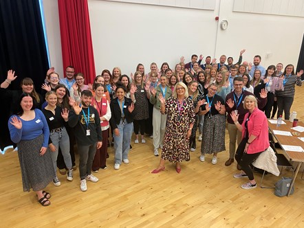 Newly Qualified Teachers starting in Moray schools this year stand in a crowd with Vivienne Cross, Head of Education, in the middle - they are all waving at the camera. They are in a school hall with a clock on the wall behind, a red curtain is open and a table with paper and water bottles is front 