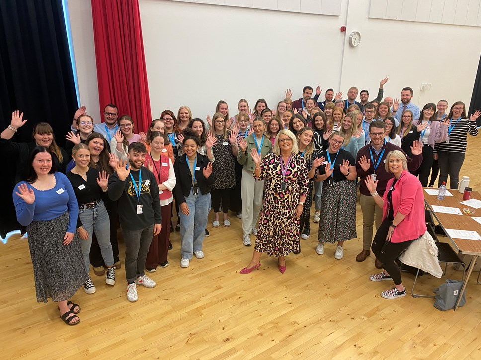 Newly Qualified Teachers starting in Moray schools this year stand in a crowd with Vivienne Cross, Head of Education, in the middle - they are all waving at the camera. They are in a school hall with a clock on the wall behind, a red curtain is open and a table with paper and water bottles is front bottom right.