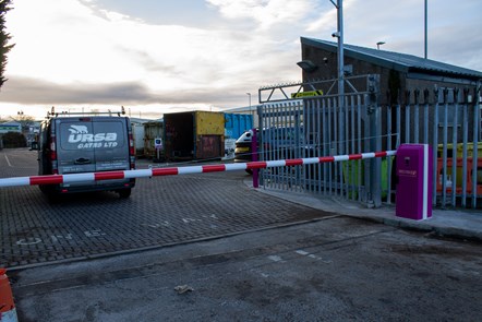 Red and white automatic barrier next to gate of Chanonry HWRC with recycling centre in the background.