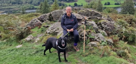 Katie's wearing a navy top, leggings and walking boots sitting on a rock with a black dog. Behind her are more rocks, a river and trees.