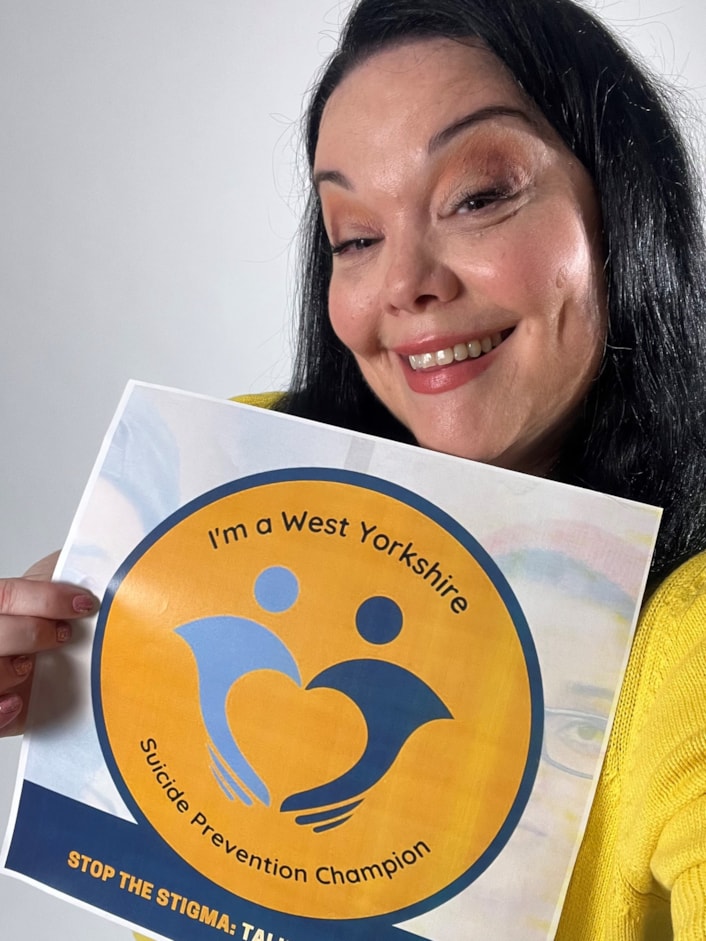 Suicide Prevention Champions - Lisa Riley A: Lisa is pictured smiling at the camera, holding the Suicide Prevention Champion logo