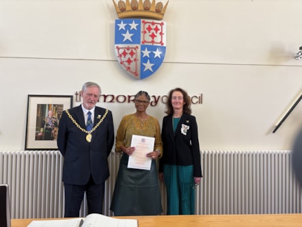 Three people stand in a line smiling at the camera. The person in the middle holds a certificate. There is a coat of arms above them.