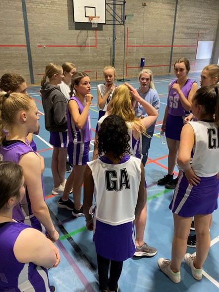 Around a dozen girls wearing netball bibs and dresses cluster around their coach for a coaching session on a sky blue-floored netball court.