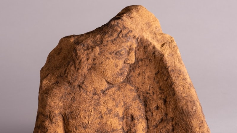 Sculpted stone panel showing a carving of a man with long curly hair, sad expression on his face and his hands tied behind his back.