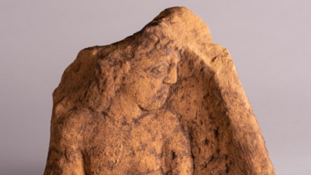 Sculpted stone panel showing a carving of a man with long curly hair, sad expression on his face and his hands tied behind his back.
