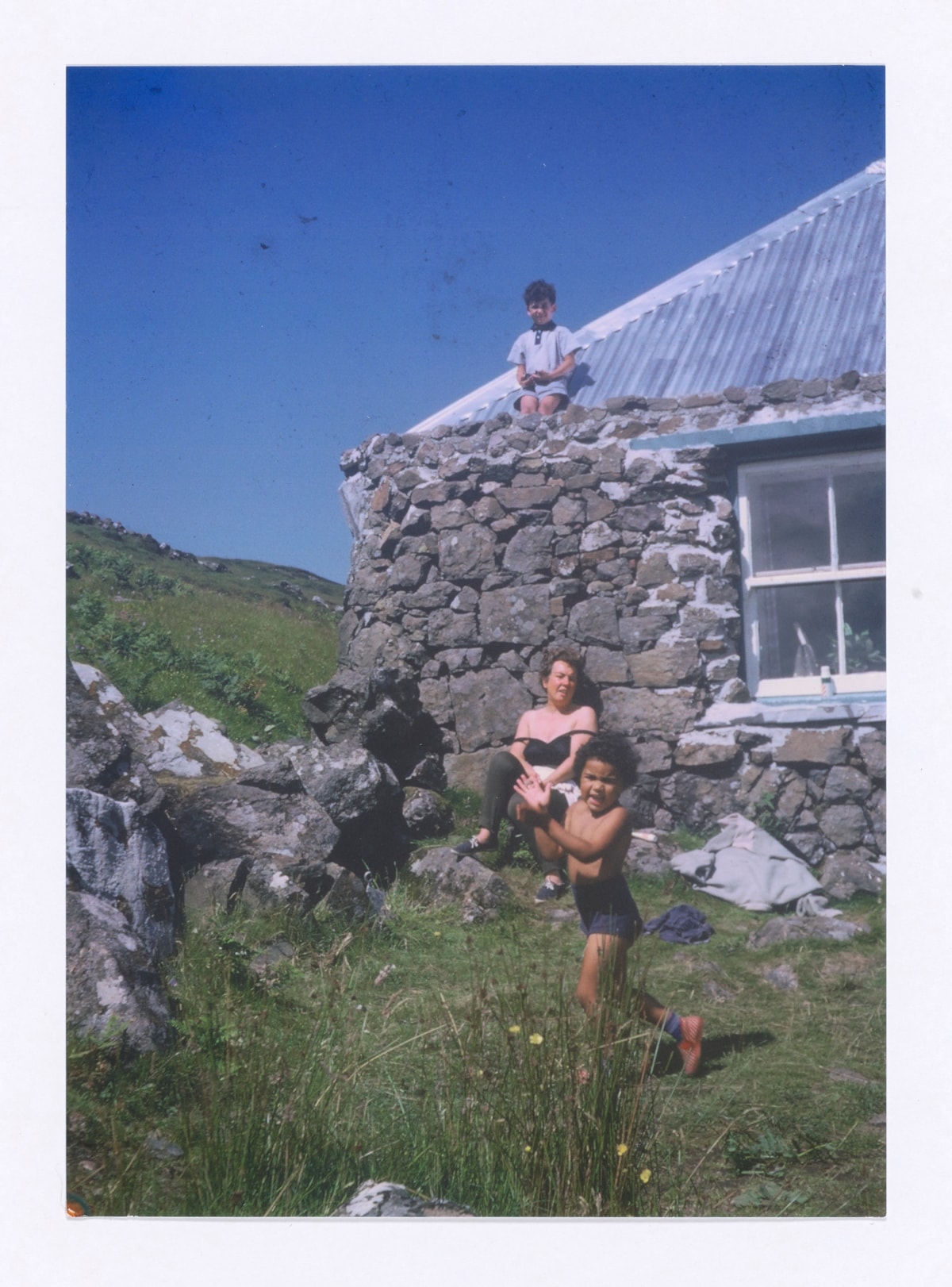 On a family holiday in Mull, Jackie Kay's mum sunbathes leaning against the croft wall. A young Jackie runs across the grass in the foreground, clapping, and her brother Maxie is perched on the corrugated metal roof.