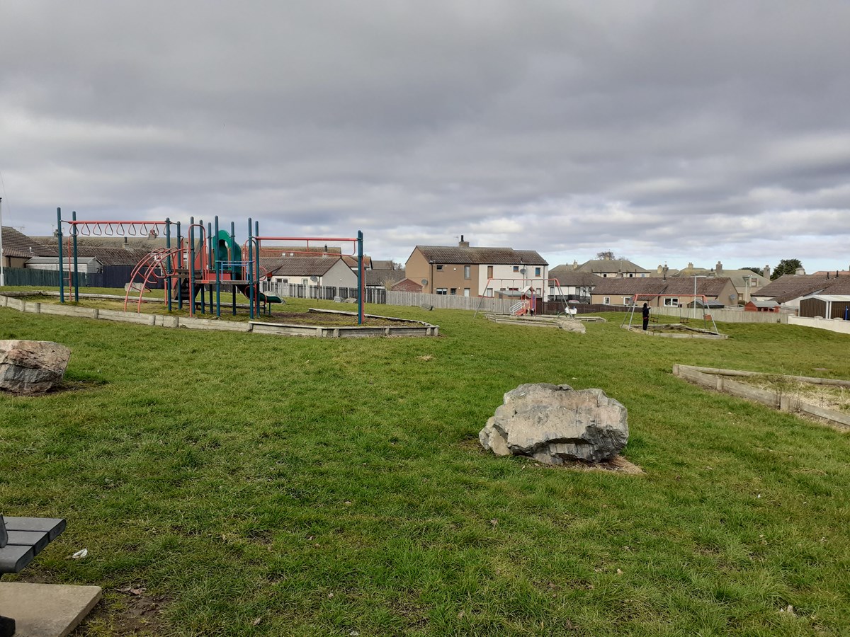 An overcast sky hangs over a small playpark, the swings and climbing frames in the distance beyond which there are several houses which dot the surrounds of the park.