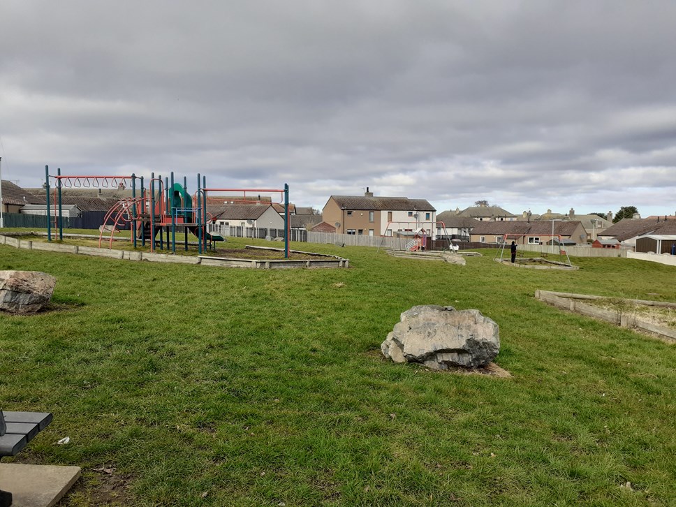 A play park in need of upgrade, there is a big grassy area and some stones and frames of swings with houses in the distance.