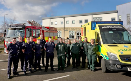 A group of fire crew and a group of ambulance crew stood together between a fire engine and ambulance in front of a  fire station building.