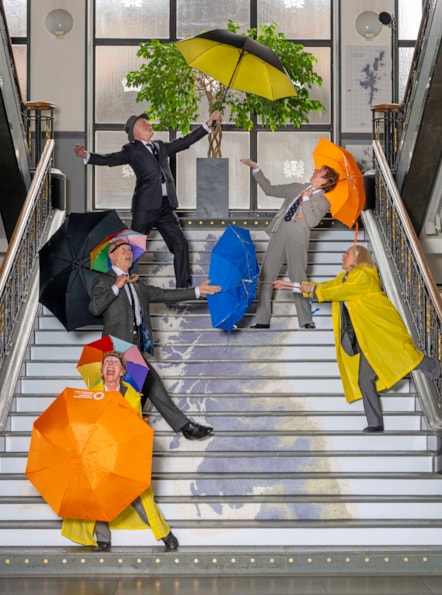 A group of dancers dressed in suits and raincoats post with colourful umbrellas on a set of stairs which has a map of Scotland printed on it