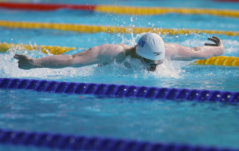 Swimmer Tom Beeley in action, doing the butterfly stroke in a swimming pool split into lanes. He is wearing a white swimming cap with "Sco" written on it.