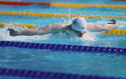 Swimmer Tom Beeley in action, doing the butterfly stroke in a swimming pool split into lanes. He is wearing a white swimming cap with 