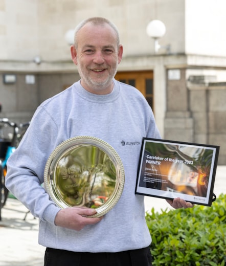 A man wearing a grey Islington Council jumper smiles and holds a winners plate and certificate outside of Islington Town Hall