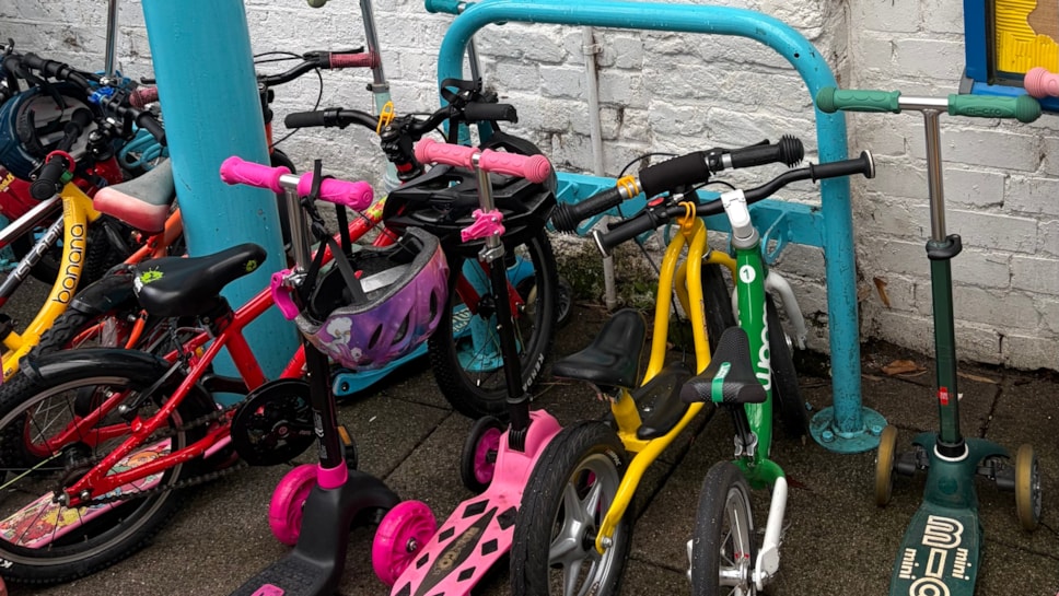 A row of colorful children's bicycles and scooters parked against a white brick wall and a blue metal rack