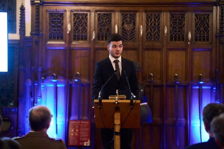 A young man aged 17 stands at a lecturn on a stage before an audience.