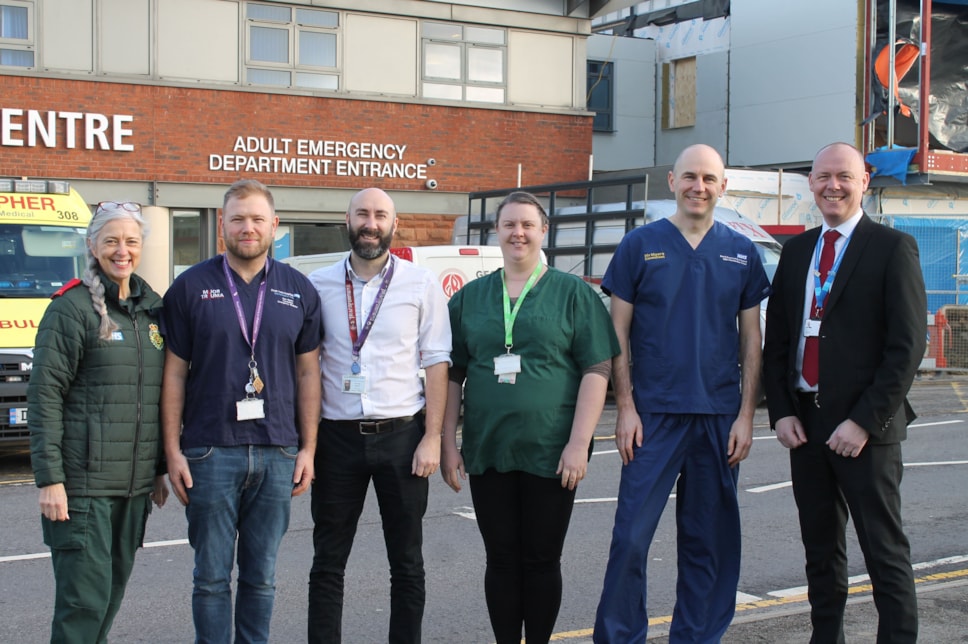 Six people who work for different parts of the NHS stood in their respective work attire in front an Adult Emergency Department