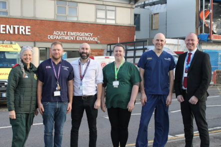 Six people who work for different parts of the NHS stood in their respective work attire in front an Adult Emergency Department