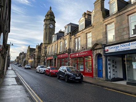 Four cars are lined up outside shops on a street with a town clock behind. There are yellow no parking lines down either side of the road.