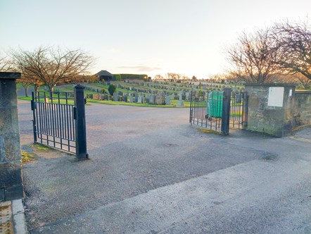 A set of grey gates are open at the edge of a cemetery, there are small trees in view and the headstones are in rows. The sky 
 is grey.