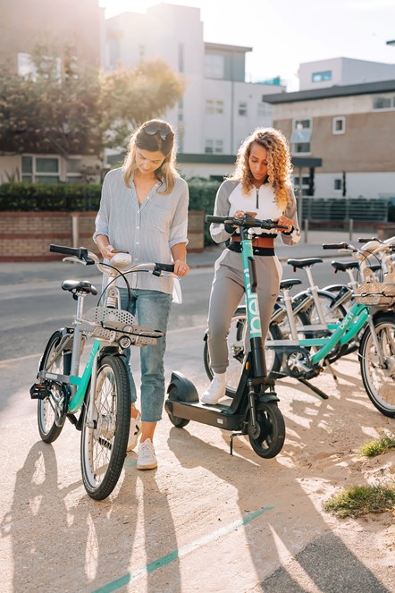 Beryl-BCP-9: Two young ladies using the Beryl app to unlock their bike (left of the picture) and e-scooter (right of the picture). Behind them is  beryl bay with other bikes and e-scooters parked up.