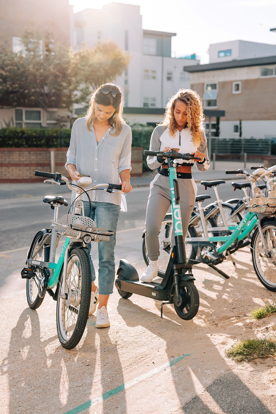 Two young ladies using the Beryl app to unlock their bike (left of the picture) and e-scooter (right of the picture). Behind them is  beryl bay with other bikes and e-scooters parked up.