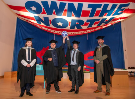 Four males in academic robes with two in the middle holding aloft a football and stood underneath a huge flag emblazoned with Own The North