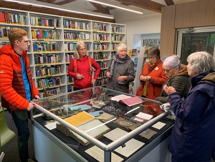 Male student with group of five females standing around an exhibition display case at the University of Cumbria Ambleside campus library
