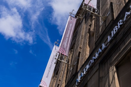 The facade of the National Library of Scotland's George IV Bridge building in Edinburgh. Two banners say 'Leabharlann Naiseanta na h-Alba and National Library of Scotland on burgundy and pale pink background respectively. Blue sky background with wispy clouds.