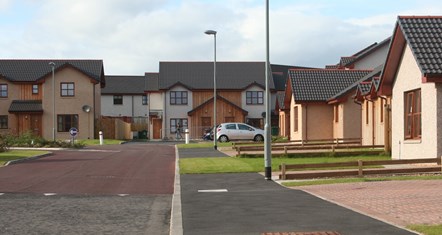 A cul-de-sac with two-story houses. A silver car is parked in a driveway and someone can be seen in the far distance walking along the road.