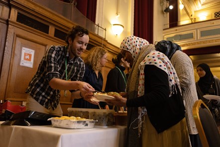 A man in a shirt and green, Islington Council lanyard serves food to a woman wearing a colourful hijab