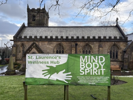 Church building with white and green sign in front that reads St Laurence's Wellness Hub, Mind Body Spirit. Light snow on the ground