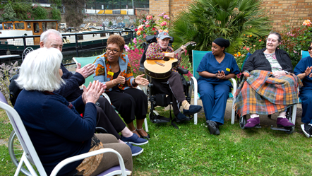 A group of smiling people sit in a garden by a canal, one plays guitar while the others clap