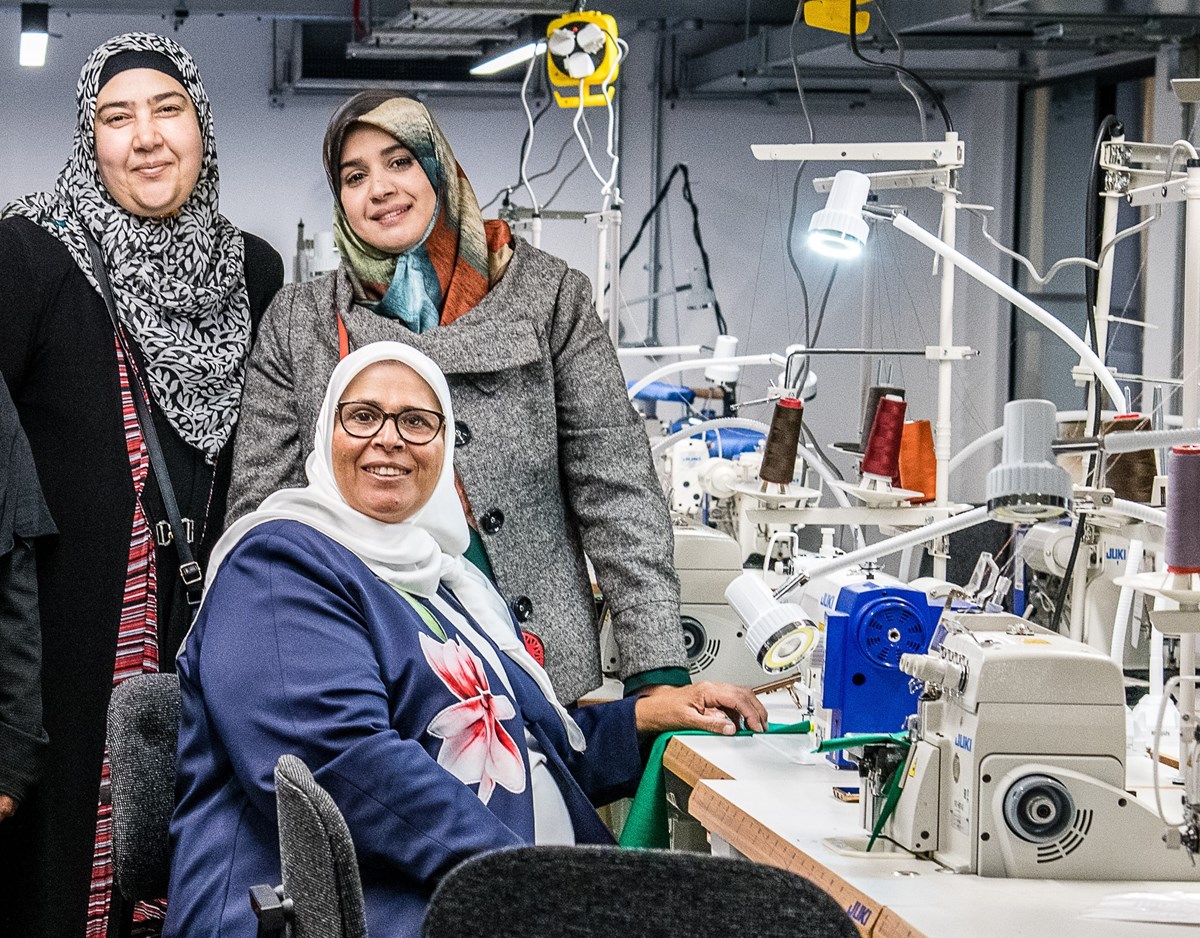 Designer-maker Majida Sayam, sitting at the sewing machine in the new FC Designer Workspace, with members of the Jannaty sewing group.