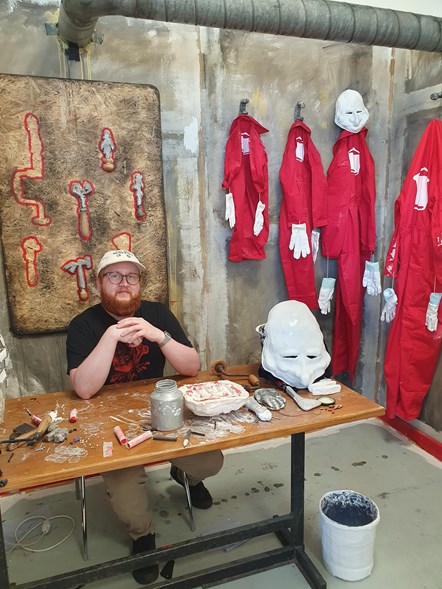 Man sitting at a table inside what appears to be an industrial  workshop with red boiler suits on hanging on the wall