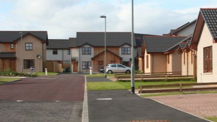 A housing scheme with a small silver car at the end of the street, there are lamp posts in the distance and a blue arrow traffic sign.