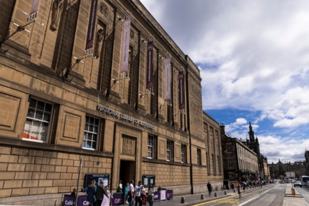 The facade of a large building on a city street. People congregating outside or walking past.