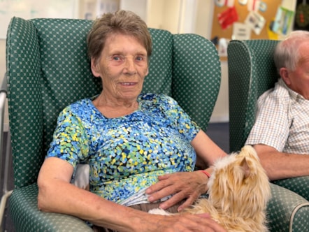 Lady wearing t-shirt with blue, yellow and green pattern is sat on a chair with a ginger and white robotic cat on her knee