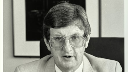 Llun du a gwyn yn dangos Colin Ford mewn siwt yn eistedd ar gadair mewn swyddfa.