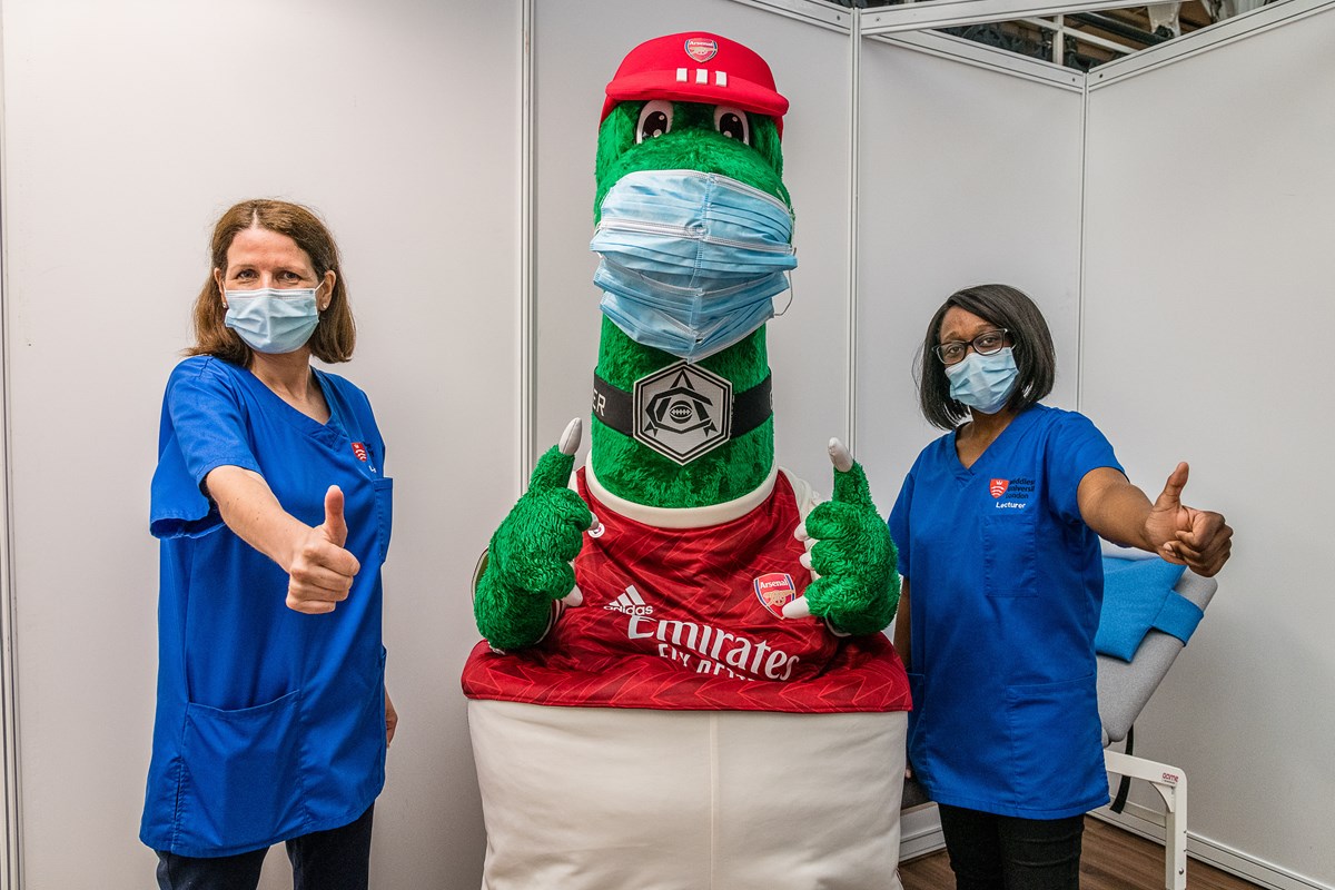 Arsenal mascot Gunnersaurus (centre), flanked by two frontline staff at an Islington vaccination centre