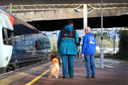 A guide dog stood on a platform alongside a train with two people next to dog's right hand side talking