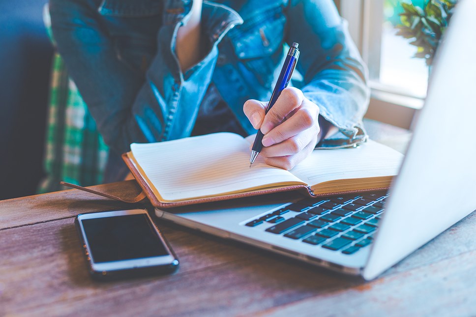 Person writing in note book with laptop and phone